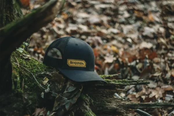 Angled View of Benjamin Trucker Hat Black BENH01 Lifestyle Outdoors Sitting on a Log in the Woods